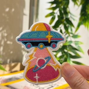 Op de afbeelding: Geborduurde patch met een kleurrijk UFO-ontwerp. De UFO is afgebeeld in blauw, geel, roze en rood, met een witte rand. De patch heeft een gedetailleerd, gestikt uiterlijk, geschikt voor toepassing op kleding of accessoires.
