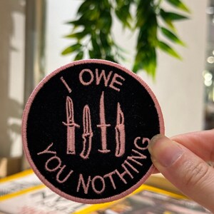 Può includere: Una toppa rotonda nera con testo ricamato rosa e bordo rosa. Il testo recita "I OWE YOU NOTHING" e presenta quattro illustrazioni di coltelli rosa.