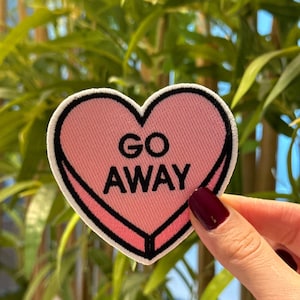 May include: A pink heart-shaped patch with the words "GO AWAY" in black. The patch has a white border and a black outline. The patch is held up in front of a green leafy background.