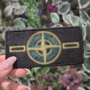Puede incluir: Un parche rectangular negro con un emblema circular que presenta un diseño similar a una brújula en verde azulado y amarillo. El parche tiene dos barras horizontales amarillas a cada lado del emblema. El parche se sostiene en una mano.