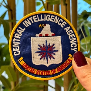 Puede incluir: Un parche circular con las palabras "CENTRAL INTELLIGENCE AGENCY" en texto blanco sobre un fondo azul marino. El parche presenta un águila blanca sobre un escudo con un diseño de estrella roja. Las palabras "UNITED STATES OF AMERICA" están en amarillo.
