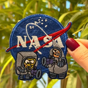 Op de afbeelding: Een blauwe, geborduurde patch met het NASA-logo en een rode boog. Twee cartoon-astronauten zijn afgebeeld, een met een hoekige helm en de andere met een ronde helm, beide met straalpistolen. Op een groene achtergrond.