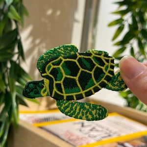 Puede incluir: Parche bordado de una tortuga marina. El parche presenta una combinación de colores verde y amarillo con un diseño detallado. El caparazón de la tortuga tiene un patrón geométrico y los bordes están delineados en amarillo.