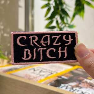 Puede incluir: Un parche rectangular negro con texto bordado en rosa que dice "CRAZY BITCH". El parche tiene un borde rosa. El parche se sostiene frente a un fondo borroso. El parche es probablemente para ropa o accesorios.