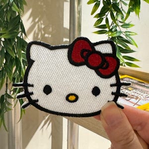 Op de afbeelding: Een wit geborduurd Hello Kitty-patch met een rode strik, zwarte ogen en snorharen en een gele neus. De patch is zwart omlijnd. Een populair karakter.