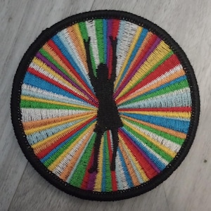 May include: Circular embroidered patch featuring a black silhouette of a person with arms raised, set against a vibrant, multicolored radial design. The patch has a black border and is likely for clothing or accessories.
