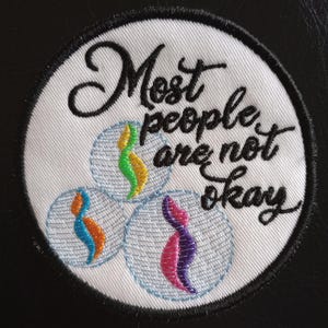 May include: Round white embroidered patch with the text "Most people are not okay" in black script. The patch features three small, stylized figures in blue/orange, green/yellow, and pink/purple within light blue circles.