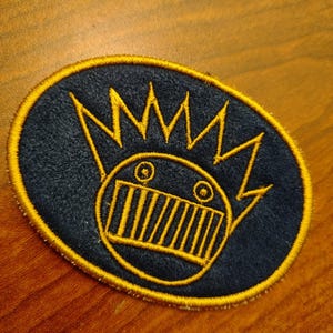 May include: An oval-shaped patch with a navy blue background and a yellow embroidered design. The design features a stylized face with a crown. The patch has a yellow border.
