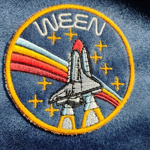May include: Embroidered patch featuring a space shuttle design with the word "WEEN" in white lettering. The patch has a blue background with a yellow border, colorful rainbow streaks, and gold star accents.