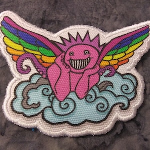 May include: Embroidered patch featuring a pink creature with rainbow wings, a crown, and a wide grin. The creature rests on a blue cloud with swirling details. The patch has a white border.
