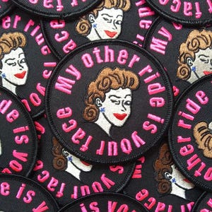 May include: A circular black embroidered patch featuring a stylized illustration of a person with brown hair and red lips. The text "My other ride is your face" is stitched in pink thread around the central figure in a circular pattern.