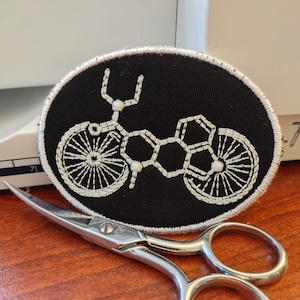 May include: An oval black patch with a white embroidered bicycle design. The patch is next to a pair of small, silver scissors on a wooden surface. The bicycle design features detailed wheels and frame.