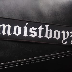 May include: Black rectangular patch with the embroidered text "moistboyz" in a gothic font. The patch is set against a textured black surface, with white stitching visible.