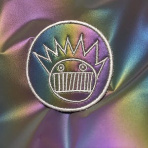 May include: A circular patch with a cartoon face design, featuring a crown and a wide-toothed grin, is embroidered with white thread. The patch has a rainbow gradient color scheme and is set against a shimmering, iridescent background.