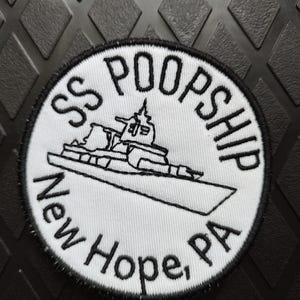 May include: A white, circular patch with a black border. The patch features the text "SS POOPSHIP" above a line drawing of a ship. Below the ship is the text "New Hope, PA" in black lettering. The patch is on a black, textured surface.