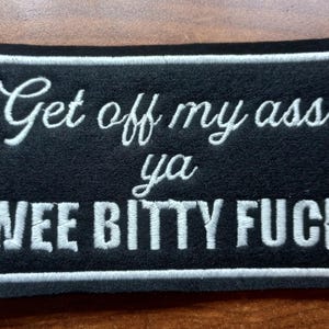 May include: Black rectangular patch with white embroidered text that reads "Get off my ass ya WEE BITTY FUCK". The patch has a white border and is on a wooden surface.