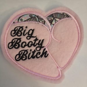 May include: A pink heart-shaped patch with the words "Big Booty Bitch" embroidered in black script. The patch has a light pink border and a patterned design at the top of the heart.