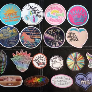 May include: A collection of twenty assorted vinyl stickers featuring various graphic designs, including text-based quotes, space themes, abstract patterns, and illustrations. The stickers vary in shape, including circles, hearts, and custom die-cut forms.
