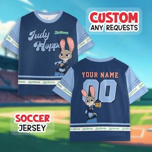 Puede incluir: Camiseta de f&uacute;tbol azul marino con Judy Hopps de Zootopia. La parte delantera muestra su nombre e imagen, mientras que la parte trasera tiene un nombre y n&uacute;mero personalizables. Mangas azul claro con texto de Zootopia. El texto "CUSTOM ANY REQUESTS" tambi&eacute;n es visible.