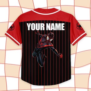 Pode incluir: Camisola de baseball vermelha e preta com acabamento branco e o texto "YOUR NAME" nas costas. A camisola apresenta um gr&aacute;fico de um super-her&oacute;i em um traje preto e vermelho. Logotipo do Homem-Aranha nas mangas.