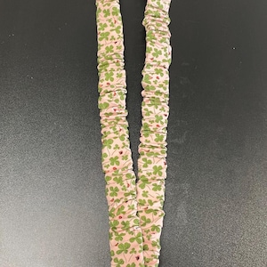 May include: A pink lanyard with a repeating pattern of green shamrocks and small red ladybugs. The lanyard is made of fabric and has a silver-colored metal clasp at the bottom for attaching keys or an ID badge.