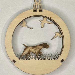 May include: A circular light-colored wooden ornament featuring a laser-cut design of a hunting dog standing in tall grass with three birds flying in the sky above. The ornament includes a top loop for hanging and has a layered, dimensional appearance.