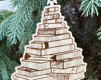 May include: Wooden Christmas tree ornament shaped like a stack of books. The ornament is made of light brown wood and features detailed engravings of book spines and pages. A star-shaped cutout tops the ornament, with a ribbon for hanging.