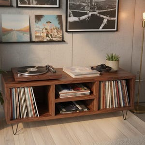 Puede incluir: Consola de tocadiscos de estilo moderno de mediados de siglo hecha de madera oscura. La consola cuenta con un tocadiscos, almacenamiento para discos de vinilo y un estante para álbumes. La consola mide 122 cm de largo y tiene patas de horquilla negras. Arte enmarcado y una planta en maceta en el fondo.