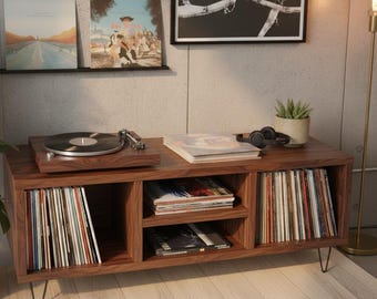 May include: A dark wood record player console in a mid-century modern style. The console has a turntable, storage for vinyl records, and a shelf for albums. The console is 122 cm long and has black hairpin legs. Framed art and a potted plant are in the background.