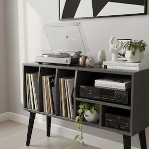 May include: A gray record player with a clear cover sits atop a dark gray media console. The console has multiple compartments filled with vinyl records, audio equipment, and decorative items, including a small potted plant and headphones.