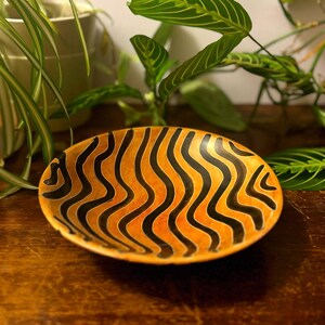 May include: Oval-shaped decorative bowl with a vibrant orange and black wavy pattern. The bowl's design features alternating black and orange stripes, creating a striking visual effect. The bowl is likely made of wood or a similar material.