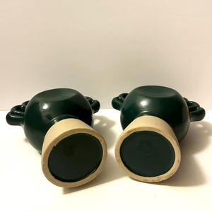 May include: Two dark green ceramic pots with cream-colored bases. Each pot has two loop handles and a circular base. The pots are displayed against a white background.