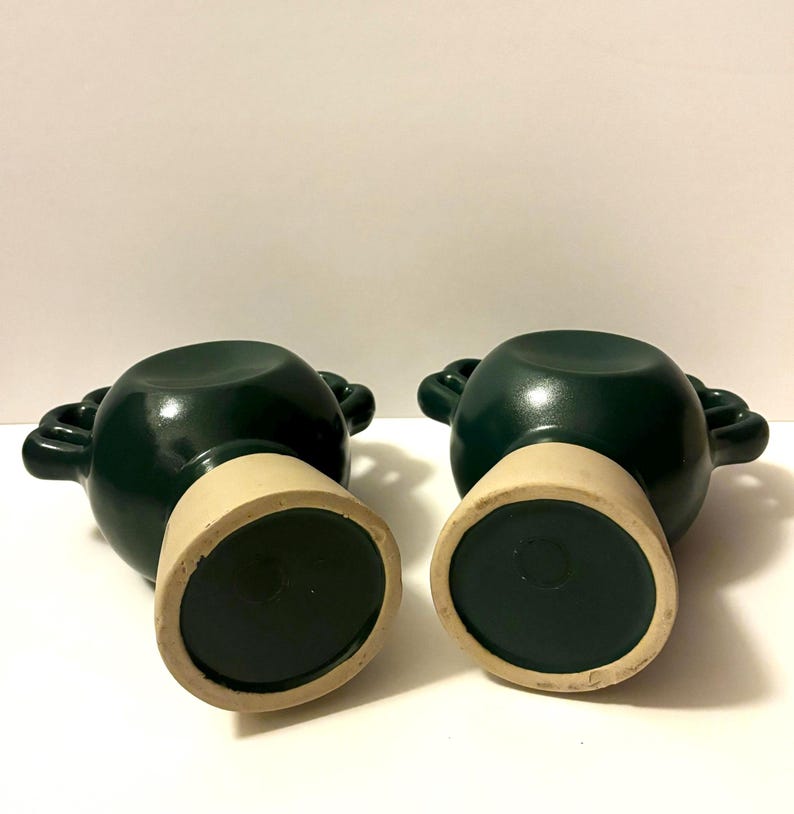 May include: Two dark green ceramic pots with cream-colored bases. Each pot has two loop handles and a circular base. The pots are displayed against a white background.