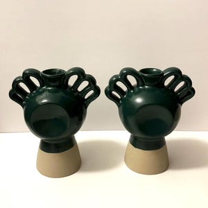 May include: Two dark green ceramic vases with a unique design. Each vase has a rounded top with arched openings and a beige base. The vases are displayed against a white background.