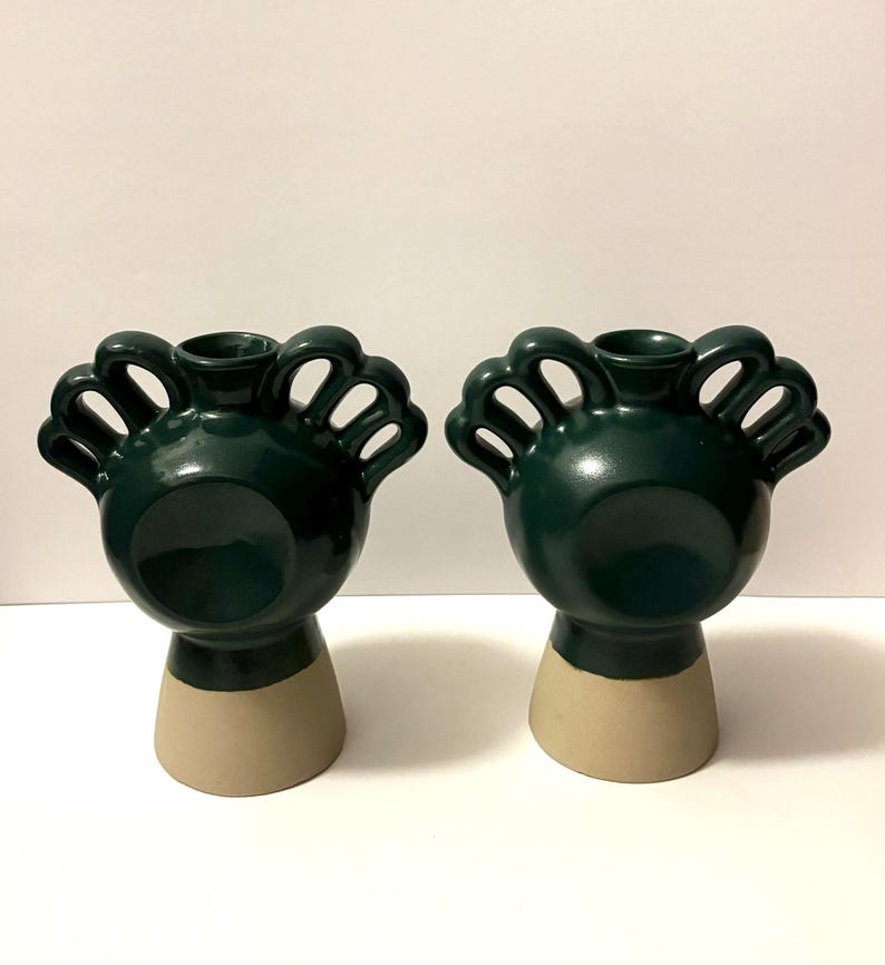 May include: Two dark green ceramic vases with a unique design. Each vase has a rounded top with arched openings and a beige base. The vases are displayed against a white background.