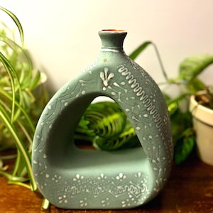 May include: A sage green ceramic vase with a unique, open design. The vase features white, intricate floral and geometric patterns. The interior is visible through the opening. The vase is on a wooden surface.