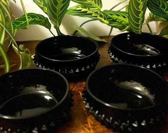 May include: Four glossy black bowls with a spiked rim design are arranged on a wooden surface. The bowls are round and have a smooth interior. Green leafy plants are visible in the background, adding a natural element to the composition.