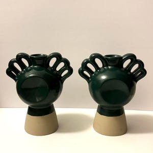 May include: Two dark green ceramic vases with a unique design. Each vase has a rounded body, a beige base, and a decorative top with open, curved shapes. The vases are displayed against a plain white background.