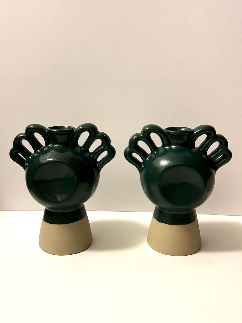 May include: Two dark green ceramic vases with a unique design. Each vase has a rounded body, a beige base, and a decorative top with open, curved shapes. The vases are displayed against a plain white background.