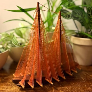 May include: A decorative wooden model boat featuring two masts with vibrant orange thread rigging stretched between the masts and the hull. The dark brown wooden hull is elongated and supports the vertical masts, creating a sail-like structure.