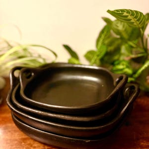 Könnte beinhalten: Ein Stapel von vier kleinen, schwarzen, oval geformten Keramikschalen mit Griffen. Die Schalen sind übereinander gestapelt, wobei die oberste Schale im Fokus steht. Der Hintergrund umfasst eine Holzoberfläche und grüne Pflanzen.