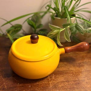 Op de afbeelding: Een felgele geëmailleerde metalen fonduepan met bijpassend deksel. De pan heeft een gladde, ronde vorm, een enkele lange houten handgreep aan de zijkant en een kleine, ronde houten knop in het midden van het deksel voor het optillen.
