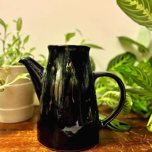 May include: A black ceramic pitcher with a curved handle and spout. The pitcher has a textured surface and sits on a wooden table. Green plants are visible in the background, adding a natural element to the scene.