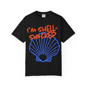 May include: A black short-sleeve t-shirt featuring a graphic of a blue seashell centered on the chest. Above the shell, the text I'M SHELL-SHOCKED is printed in a bold, orange, hand-lettered style font. The shirt has a standard crew neckline.