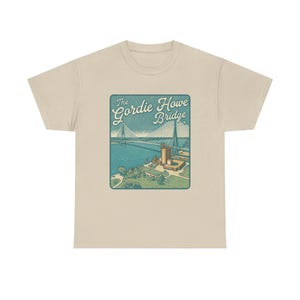 May include: A beige t-shirt featuring a vintage-style illustration of the Gordie Howe Bridge. The design includes the bridge, water, and a park, with the text "The Gordie Howe Bridge" in a teal and white color scheme.