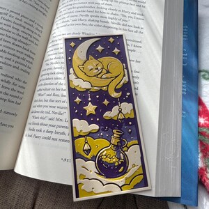 May include: A bookmark featuring a sleeping cat on a crescent moon, surrounded by stars and clouds. Below, a potion bottle with a swirling liquid is depicted. The color palette includes yellow, purple, and white, with a whimsical, illustrated style.