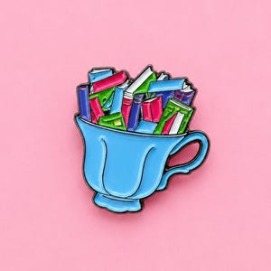 May include: A metal enamel pin shaped like a light blue teacup filled with a stack of colourful miniature books. The books are rendered in shades of red, green, blue, and purple, with white pages visible, all outlined in a thin black metal border.