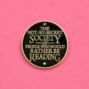 May include: A circular black enamel pin with a gold-colored metal border. The center features gold lettering that reads: THE NOT-SO-SECRET SOCIETY OF PEOPLE WHO WOULD RATHER BE READING, accented with small gold diamond shapes at the top and bottom.