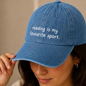 May include: A blue denim baseball cap with the embroidered text "reading is my favourite sport." The cap features a curved peak and a rounded crown. This casual hat is a classic style, perfect for everyday wear.