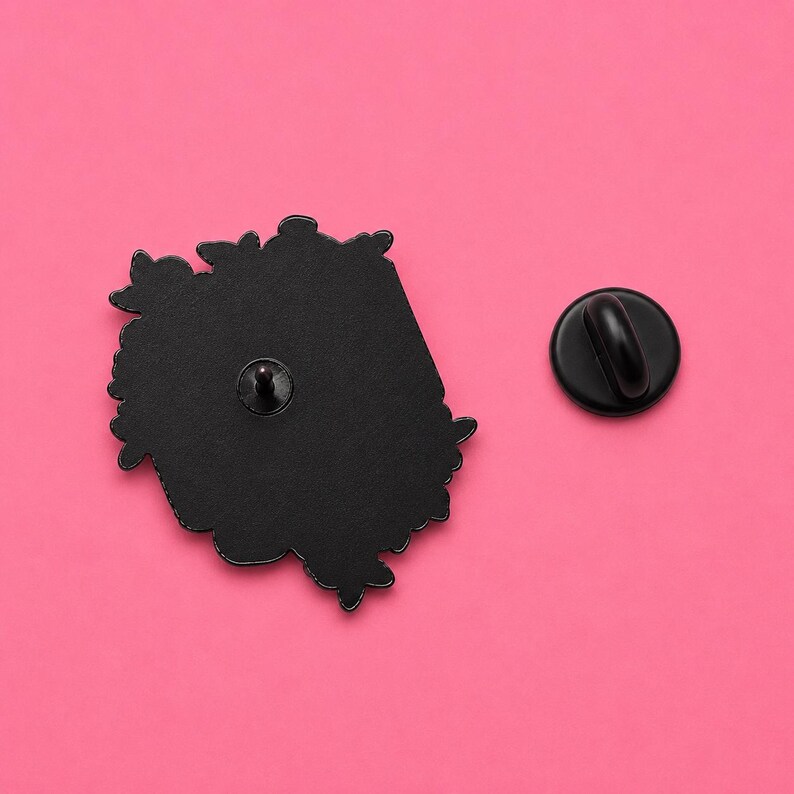May include: The reverse side of a black metal enamel pin featuring a textured, irregular floral-shaped outline. A central metal post protrudes from the back, accompanied by a separate, circular black metal clutch fastener for secure attachment.